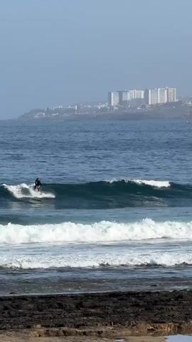 This is a video of a surfer riding a wave near Playa De Las Américas, Spain. The surfer is wearing a black wetsuit and is positioned on a white surfboard. They are riding a small, breaking wave, with white foam visible around the board. In the background, there are several large, white buildings, likely hotels or apartments, situated on a hill overlooking the ocean. The sea is a deep blue, with multiple waves breaking in succession. The sky is hazy and overcast, suggesting it might be early morning or late afternoon. The foreground shows a rocky shoreline with dark, wet rocks. The overall scene conveys a sense of coastal activity and a relaxed atmosphere. There is no visible text in the video.