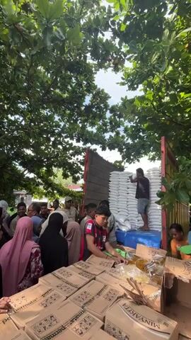 A group of people, predominantly women wearing headscarves, are gathered around a truck that is being unloaded. The truck is filled with large white sacks. In the foreground, there are stacks of cardboard boxes and some yellow-packaged items, possibly food items. Some individuals are actively engaged in handling the boxes, while others appear to be waiting or observing. The scene takes place outdoors, under the shade of large leafy trees, with sunlight filtering through the foliage. The overall atmosphere suggests a distribution of goods, possibly for aid or a community event. The video pans to show a longer line of people, many also wearing headscarves, extending down a dirt path lined with trees and some houses in the background. The sun is bright, casting rays through the leaves, and a motorbike is parked on the side of the path.