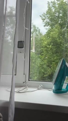 The media file shows a close-up view of a white windowsill with a white plastic window frame. On the windowsill, there is a teal and white iron with the brand name "Tefal" visible. The iron is connected to a white cord that is coiled on the windowsill. To the left of the iron, attached to the window frame, is a small digital thermometer displaying "22.1" and what appears to be a hygrometer. A white curtain is draped on the left side of the window. Outside the window, lush green trees are visible, suggesting an outdoor, natural setting. The sky appears overcast, indicating cloudy weather. A thin string of fairy lights, with small white bulbs, is draped vertically along the window frame. A traditional mercury thermometer is also attached to the windowpane on the outside, with the red liquid line visible. The scene appears to be set in a residential interior, likely a room with a view of nature. There are no people visible in the media file. The overall mood is calm and domestic, with a focus on everyday objects and the view outside. The time of day is not explicitly indicated but the lighting suggests daytime.