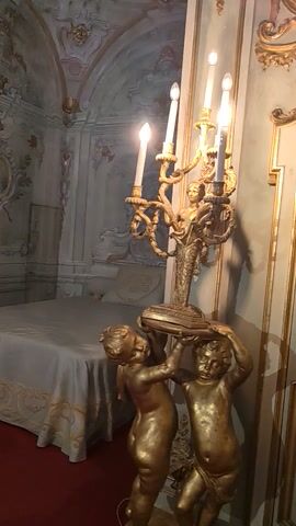 The video shows a bedroom with a bed covered in a white sheet with gold trim. The room has ornate molding with gold accents on the walls and ceiling. There is a chandelier with lit candles visible in the foreground and a statue of two figures above the bed. The floor is red. The camera appears to be positioned on the floor and rotated to display the ceiling.