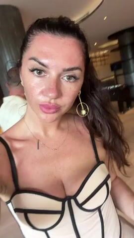 The video shows a woman taking a selfie in what appears to be a modern hotel lobby or lounge area. She is wearing a light-colored, strapless, corset-style top with black trim and large, circular earrings with a starburst design. Her hair is dark and pulled back, and she has on makeup, including lip gloss. In the background, a man in a patterned shirt and shorts walks away from the camera, carrying a black bag. The environment features contemporary furniture, including an orange sofa and a pool table in the distance, with warm lighting. The woman is looking directly at the camera and speaking, though the audio is not transcribed. She occasionally touches her hair and adjusts her posture.