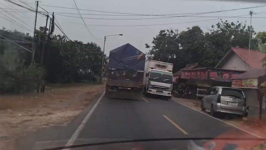 The video captures a dashcam view of a road with two trucks and several other vehicles. The first truck, a white refrigerated truck, is parked on the right side of the road. Further ahead, a large truck with a blue tarp-covered load is moving slowly in the same lane. A motorcycle passes the white truck and moves towards the large truck. On the left side of the road, a white van is visible. The road appears to be a two-lane road with a dirt shoulder on the left. The environment is rural, with trees and buildings visible on either side of the road. The sky is overcast.
