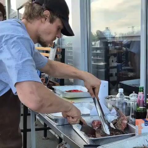 A man wearing a baseball cap, blue shirt, and brown apron is preparing food at a table. He is using tongs to hold a piece of meat in a foil pan while a knife in his other hand is positioned over a white surface in another foil pan. The table is a stainless steel surface. There are other foil pans on the table containing food and ingredients. Bottles and containers of spices and seasonings are visible on the table. Behind the man, there is a window showing reflections of people and shelves. The scene appears to be outdoors.