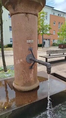 This is a close-up, medium-shot video of a water fountain in Gravenbruch, Germany. The fountain has a thick, reddish-brown stone column with a decorative top, reminiscent of a classical column. Attached to the column is a dark metal spout, shaped like a dragon's head, from which a stream of clear water is flowing into a basin. The water creates a cascade of droplets as it falls.

Behind the fountain, a modern, multi-story building with an orange and white facade is visible. Trees with green foliage line the sidewalk, and a park bench sits in the background, suggesting a public outdoor space. The sky appears cloudy, indicating an overcast or partly cloudy day.

A small sign is affixed to the fountain's column. It contains text that reads "Kein Trinkwasser," which translates to "No Drinking Water" in German. The presence of this sign is a notable detail, indicating that the water from the fountain is not safe for consumption. The overall scene appears calm and peaceful, with the focus being on the gentle flow of water. There are no people directly in the foreground or interacting with the fountain, although a person is faintly visible walking in the background on the left side of the frame.