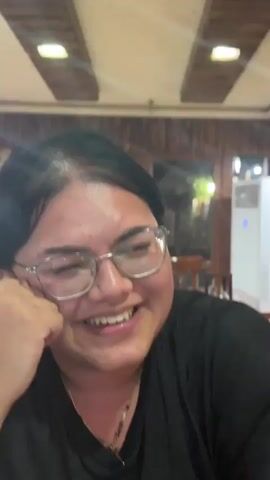 The video shows a close-up of a person wearing glasses, with dark hair and some grey streaks. They are smiling and laughing, with their eyes mostly closed at times. Their hand is resting on their cheek. The person is wearing a black shirt and several necklaces. The background appears to be indoors, possibly a restaurant, with wooden paneling on the ceiling and some blurred lights. The overall mood seems cheerful and lighthearted.