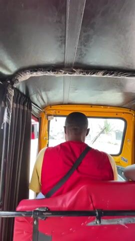 The video shows the back of a man wearing a red shirt and a black shoulder bag driving a yellow tuk-tuk. The interior of the tuk-tuk has a black roof, red seats, and gray curtains.  Outside the tuk-tuk, other vehicles and people are visible. The windshield wipers are in operation.