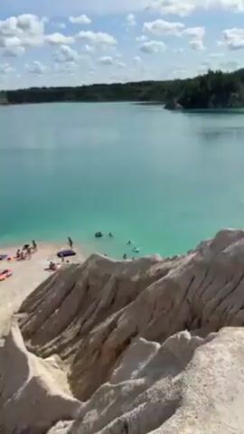 The video depicts a scenic landscape featuring a clear turquoise lake surrounded by white, rocky formations reminiscent of cliffs or dunes. The water is placid, with a small beach area visible where several people are gathered, some swimming or wading in the water. On the opposite side of the lake, a dense green forest lines the shore, providing a stark contrast to the light-colored lake and rocky terrain. The sky is partly cloudy, with patches of blue visible. The overall impression is one of natural beauty and tranquility.