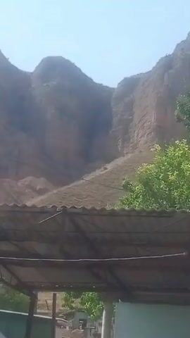 The video shows a view of a large, steep, and barren mountain or cliff face under a clear blue sky. The rock appears to be sedimentary, with visible layers and erosion patterns. In the foreground, a portion of a covered structure with a corrugated roof supported by metal posts is visible. Below this structure, there are trees with green foliage and what appears to be a wall or fence in the very bottom of the frame. The lighting suggests it is daytime, likely morning or afternoon, with the sun casting shadows and highlighting the texture of the mountain. There are no people or animals visible in the frame. The overall impression is of a natural landscape with some human-made structures in the immediate foreground.