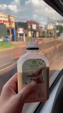 A first-person view shows a hand holding a bottle of Ricca milk tea with text reading “Tra Sua O Long Lai”. The bottle is plastic and clear with a white and light green label. The bottle is mostly empty, as it is just filled to below halfway. A person’s hand can be seen holding the bottle with a small tattoo on their wrist. The background is out of focus and shows a street with buildings and vehicles passing by the window. The video appears to be taken from inside a moving vehicle. The camera angle is slightly angled downward, showing a portion of the gray seat. The natural light and color appears normal.