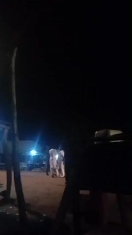 The video shows an outdoor scene at night. A bright street lamp illuminates the area. In the foreground, there is a wooden structure, possibly a shelter or stall, with tables and chairs visible underneath. To the left, a person wearing blue is sitting at one of the tables. Further in the background, several people are standing or walking around. The ground appears to be dirt or sand. The overall atmosphere suggests a gathering or activity taking place in a public space after dark.