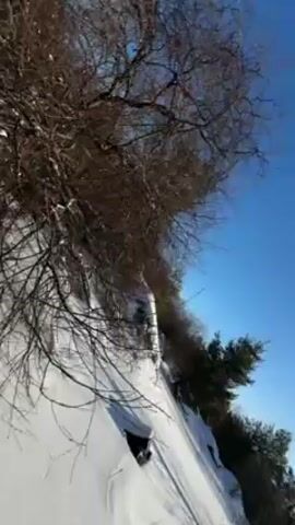 A woman with long brown hair wearing a black jacket and beanie stands in a snowy landscape on a sunny day. She holds the camera at arm's length, filming herself from a low angle. As the camera moves, a large cooling tower comes into view behind her. The sky is clear and blue, and there are some bare trees surrounding her.