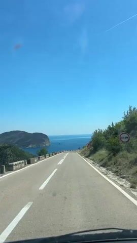 The camera is positioned inside a vehicle traveling on a two-lane road. The road is paved and has white lines marking the lanes. The road curves slightly to the left. On the left side of the road is a low stone wall. Beyond the wall is a body of water with an island in the distance. On the right side of the road is a hillside covered with green vegetation. There is a triangular road sign with a curved arrow on the right side of the road. The sky is clear and blue.