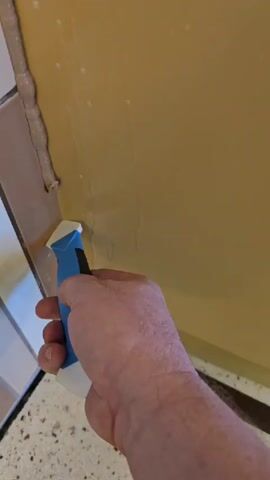 A close-up view shows a person's right hand holding a blue and white caulking tool. The tool is being used to smooth a bead of light brown or beige caulk along the joint between a tiled surface and a yellowish wall or panel. The tiled surface features square tiles in white and a lighter brown shade. The caulk is being applied neatly, forming a continuous line. Small bubbles or drips are visible on the yellowish surface, adjacent to the caulk line. The scene suggests an indoor setting, likely a bathroom or kitchen, where sealing joints is common. No text is visible.