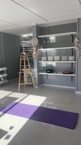 The video shows a woman with blonde hair tied in a high ponytail, wearing a black t-shirt and burgundy shorts. She is in a room with light gray walls and a white ceiling. The room appears to be a yoga or meditation studio, with a purple yoga mat laid out on the floor. In the background, there are white shelves holding several crystal singing bowls and other decorative items, including dreamcatchers. An easel stands to the left of the shelves. The woman walks into the frame from the left, faces the camera, then turns to face the yoga mat. She stands on the mat with her feet shoulder-width apart, then walks forward and turns to face the camera again with a smile. She is barefoot throughout the video. Sunlight streams into the room from the left, creating shadows on the floor and mat.