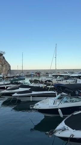 The video shows a view of a marina with several boats docked. The boats are of various sizes, with some appearing to be speedboats and others possibly sailboats. The water in the marina is calm, and the sky is clear and blue. In the background, there is a rocky cliff face with some vegetation. The overall atmosphere of the video is peaceful and serene, possibly taken at sunrise or sunset.