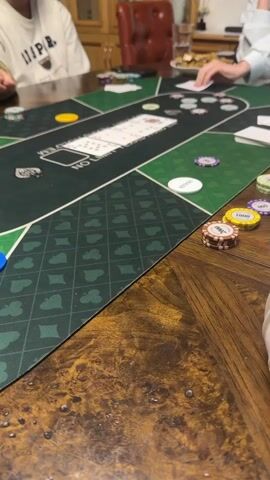 This video shows a low-angle, static shot of a poker table. The table is green with a patterned border. Several stacks of poker chips are visible on the table, in denominations of 1000 and smaller. There are also playing cards and what appear to be betting markers on the table. At least three people are partially visible, seated around the table, their hands and upper bodies in the frame. One person is wearing a white sweatshirt with text on it, another is wearing a dark-colored jacket, and a third is wearing a light-colored shirt. The environment appears to be an indoor setting, possibly a home, with a wooden table and some background furniture visible. The lighting is dim. The action appears to be a poker game in progress, with chips being moved and cards potentially being handled.