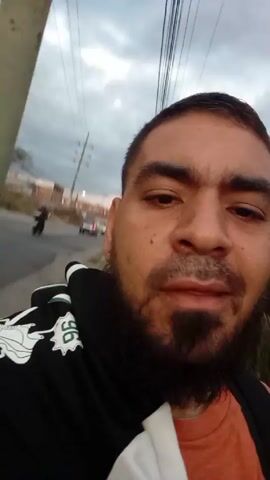 A man with a beard and mustache is standing outside on a road and recording a video with a mobile device. The man has a black and white hooded jacket on over an orange shirt. Behind him is a road, a grassy area, a utility pole and power lines, and a cloudy sky. A vehicle drives past in the background. The man smiles and speaks to the camera.