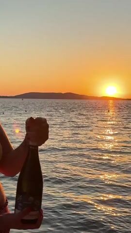 In this media file, a person's hands are visible holding a dark green bottle of what appears to be champagne or sparkling wine. The bottle is being uncorked, as evidenced by the hands twisting the foil and wire cage around the cork. The background features a vast expanse of calm, rippling water, reflecting the warm, golden hues of a sunset. A distant, low-lying island or coastline is silhouetted against the vibrant orange sky, where the sun is partially visible, casting a shimmering path across the water.

The time of day is clearly sunset, indicated by the warm, low-angled light and the intense orange and yellow colors in the sky. The weather appears to be calm and pleasant, with the water being smooth. The overall mood evoked is celebratory and serene, likely a moment of enjoyment and relaxation by the sea.

The visible text on the bottle label is partially obscured and difficult to read clearly. However, some letters and shapes can be discerned, suggesting a wine or beverage label with possibly an illustration. The location context provided, Grad Šibenik, Croatia, places this scene along the Adriatic coast, known for its beautiful sunsets and coastal landscapes.