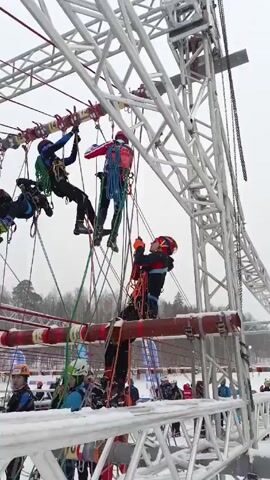 The video shows a group of people participating in an outdoor obstacle course in a snowy environment. Some participants are hanging from ropes and climbing across overhead structures while others are on the ground, seemingly preparing for their turn. The course is made of metal scaffolding with various ropes, beams, and other obstacles suspended between the structures. People are wearing helmets and safety harnesses. The background shows trees and a large screen. The overall impression is of a challenging and physically demanding activity in a winter setting.