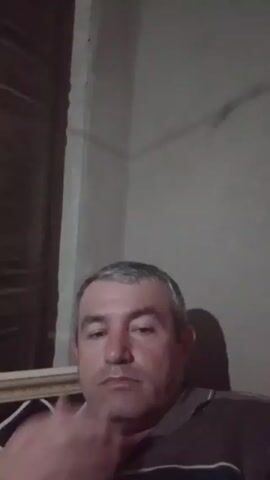 The video shows a man with graying hair in a dark room. He is wearing a dark shirt with stripes. He initially covers his face with his hand, then he gives a thumbs up. The background is a wall, part of a window, and a piece of furniture with a patterned edge. The lighting is dim, casting shadows on the wall.