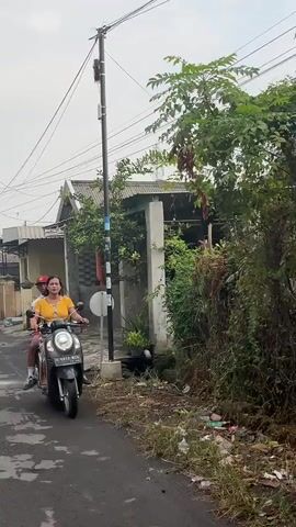 A man and a woman ride on a scooter down a paved road. The woman is in front, wearing a yellow shirt and red hat. The man is behind her, wearing a gray shirt. They are passing by houses and trees. The sky is overcast. There is some trash on the side of the road.