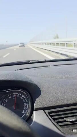 The video captures a forward-facing view from the interior of a moving vehicle on a multi-lane highway or bridge. The dashboard and a portion of the steering wheel are visible in the foreground, with a speedometer showing a speed of approximately 100 km/h. A gray speaker grille is prominent on the right side of the dashboard. Ahead, the road stretches out with a white car visible in the distance. To the right, a long metal barrier or sound wall runs parallel to the road, with vertical support poles. The sky above is clear and blue. The scene is brightly lit by sunlight.
