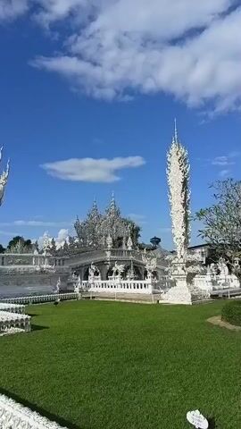 A video captures a series of sweeping shots of a white temple against a bright blue sky with scattered clouds. The temple's intricate white architecture is detailed with spires, ornate carvings, and decorative elements. The camera pans across the grounds, revealing a well-maintained green lawn and pathways leading to the temple entrance. The scene is bathed in sunlight, highlighting the pristine white facade of the structure.