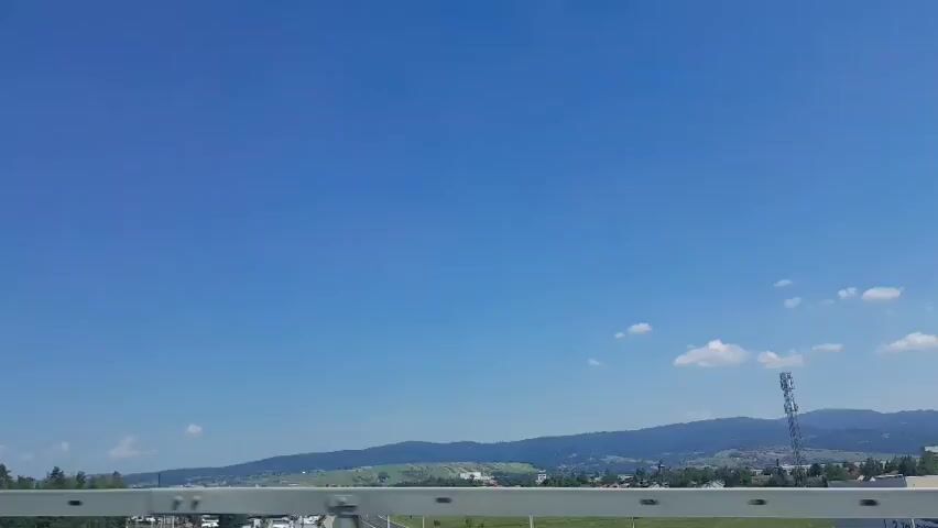The video captures a scenic view from a moving vehicle, likely a car or train, on a bright, clear day. The sky is a vibrant, cloudless blue, suggesting fair weather. In the foreground, a silver railing runs horizontally across the bottom of the frame, indicating the viewer is on a bridge or elevated structure. To the right, a white pole with a red and orange band is visible, possibly a light pole or a marker.

The middle ground reveals a landscape of rolling green hills and mountains in the distance. A highway or road with visible traffic snakes through the valley below, bordered by green fields and scattered trees. Some buildings and industrial structures are discernible in the valley, hinting at a town or city in the vicinity. The overall setting evokes a sense of travel through a rural or semi-rural area on a pleasant day.

There are no visible people in the frame. The activity taking place is implied to be travel, with the camera perspective moving forward, offering a dynamic view of the passing scenery. The time of day is clearly daytime, given the bright sunlight and clear sky. The location context of Nowy Targ, Poland, places the scene in a region known for its natural beauty, which is consistent with the rolling hills and mountainous backdrop. No text is clearly visible within the footage, though some indistinct markings might be present on the white pole. The overall mood conveyed is peaceful and expansive, with the vast blue sky and distant landscapes offering a sense of freedom and exploration.
