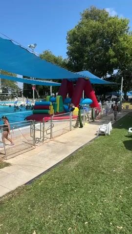 A bright sunny day at a public pool. A large, inflatable water obstacle course is set up near the edge of the pool. The obstacle course is red, blue, green, and yellow, and it features climbing sections, a slide, and a ramp. People are swimming and playing in the pool. Some people are standing on the deck near the obstacle course. There are lounge chairs and umbrellas set up on the grass. The overall atmosphere is one of recreation and fun.