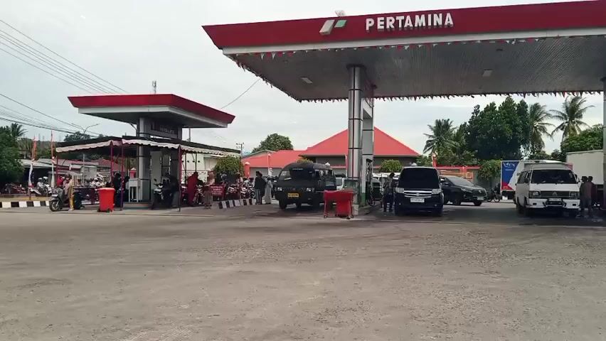 The video shows a busy Pertamina gas station. The main structure is a large red and white awning with Pertamina branding. There are multiple cars and motorcycles at the pumps. People are seen refueling their vehicles and interacting at the service area. A small building with a red roof is visible behind the pumps, with some people standing near it. The weather appears to be overcast. The ground is paved and dry.