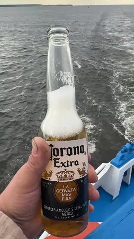 The video begins with a close-up shot of a hand holding a bottle of Corona Extra beer with foam spilling over the top. The background is a blurry body of water. The camera then pans away from the bottle to show the wake of a boat moving through the water. A fishing net is visible trailing behind the boat, catching some debris in the water. The video continues to show the churning water and the wake of the boat as it moves further away. The scene appears to be on a ferry or a similar vessel, with a glimpse of the boat's railing. The sky is overcast and the water is choppy.