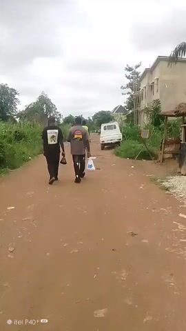 This video shows a dirt road in Achara Ndiagu Omegio, Nigeria. Two men, seen from behind, are walking away from the camera down the road. The man on the left wears a black t-shirt with a graphic design and black pants. The man on the right wears a grey t-shirt and black pants, carrying a white plastic bag. A third person in a yellow shirt is visible further down the road, and a white pickup truck is parked to the right. Lush green vegetation lines both sides of the road, with a partially constructed building visible on the right. The sky is overcast with heavy clouds, suggesting it might be a cloudy day or perhaps evening. The ground is uneven and appears dusty, with some scattered litter. The overall atmosphere is ordinary and unremarkable, depicting everyday life in a rural or semi-rural setting. The timestamp at the bottom left indicates "itel P40+".