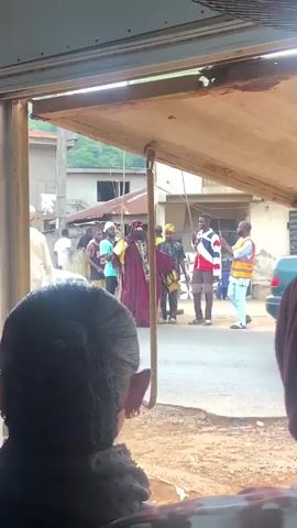 Here is a description of the video:
A video shows a cultural performance on a street, viewed from inside a building. The perspective is through a window or opening, framing the scene outside. In the foreground, the back of two people's heads are visible, slightly obscuring the view. A person dressed in traditional attire with red and gold striped clothing and a headdress is visible in the center of the frame. This person is holding a golden object. Two men stand to the side, one in a red, white, and blue striped jacket and the other in a safety vest and shirt. Additional people can be seen in the background, along with houses and cars, creating a neighborhood scene. The video is shot at eye level, with the viewer seemingly an observer from within a building.