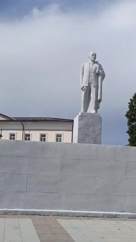 This video shows a statue of Vladimir Lenin in a public square in Irbit, Russia. The statue is made of white stone and is mounted on a pedestal. In the background, there are several buildings, including a brick building that appears to be in disrepair. There are also trees and a cloudy sky. A woman, partially visible, is walking in the foreground. She is wearing a pink jacket and has a white hair clip. The overall impression is of a slightly overcast day in an urban setting. The video appears to be taken in the daytime. There is no visible text in the video.