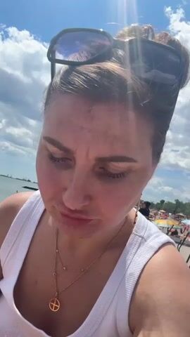 The video shows a close-up of a woman's face as she eats ice cream with a spoon. She is wearing a white tank top and sunglasses are perched on her head. The background suggests an outdoor setting, possibly a beach, with a blue sky and scattered clouds. There are other people and beach umbrellas visible in the distance. The woman's expression shifts from looking away to looking at the camera with a slight smile as she eats.