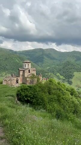 The video opens with a view of an ancient stone church situated on a grassy hill. The church has a domed roof and appears to be partially ruined. Several people are visible standing near the church, possibly tourists. The background is dominated by rolling green hills covered in lush vegetation, with a cloudy sky above. The camera then pans to the right, revealing a wide valley with a river winding through it. More forested hills stretch into the distance, and the sky remains cloudy. The foreground consists of tall green grass with some small wildflowers. The overall environment is natural and scenic, with the ancient church as a prominent focal point.