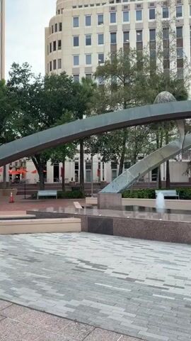 The video shows an outdoor urban scene with a fountain in the foreground and buildings in the background. The fountain has a modern design with a metal archway and water jets. The ground is paved with interlocking gray and beige bricks. To the right of the fountain, there is a silver park bench. Behind the fountain, there is lush green grass and several trees, including palm trees. In the background, there are multi-story office buildings with many windows. One building is lighter in color and has a curved facade, while another is darker and has a more angular design with a glass exterior. A bright green electric scooter, a Lime bike, is parked on the paved area to the right of the fountain and in front of the palm trees. In the distance, two people are visible sitting on benches near the fountain. The sky appears overcast.