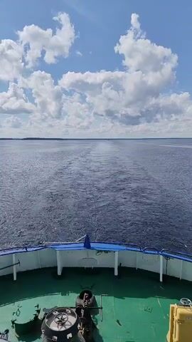 The video shows the wake of a boat as it moves through a large body of water. The perspective is from the stern of the boat, looking back. The water is dark blue and choppy, with a distinct V-shaped wake stretching out behind the boat. The sky is blue with scattered white clouds. The deck of the boat is visible in the foreground, with some equipment and railings. There are no people visible in the video.