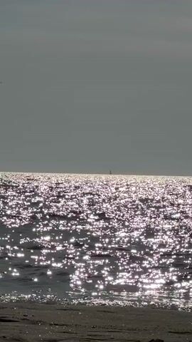 The video shows a calm sea with sunlight reflecting off the surface, creating a sparkling effect. The sky is overcast, and the horizon is visible in the distance with a small sailboat. The foreground shows a sandy beach with some small pebbles and debris. The camera is still.