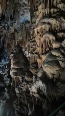 The video shows the interior of a cave. It features numerous stalactites and stalagmites of varying sizes and colors, ranging from dark browns to lighter tans. The cave's rock formations are illuminated by strategically placed lighting fixtures on the ground. The camera pans upwards, revealing more of the cave's ceiling and intricate rock structures. The scene appears to be a tourist attraction, designed to showcase the geological features of the cave.