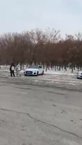 The video shows an outdoor scene on what appears to be a road or wide paved area, potentially near a university or institutional building, on an overcast day with light snow or slush present. The camera initially focuses on two people walking and a white car parked to the side of the road. The camera then pans left to reveal a long building with numerous windows and what seems to be several parked cars in the distance, including a dark-colored sedan passing by. Bare trees line the area, and the overall atmosphere appears cold and quiet.