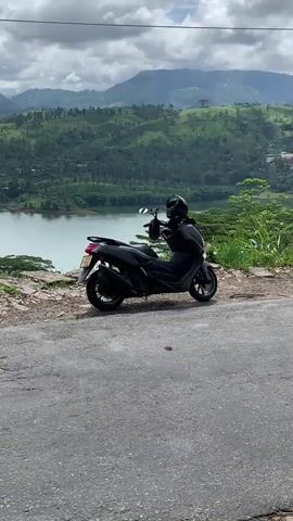 A black scooter with a helmet on the handlebars is parked on the side of a paved road. The scooter is facing to the left and is positioned in the foreground of the image. Behind the scooter, a large body of water, likely a lake or reservoir, stretches across the frame. The water is calm and reflects the sky. The background consists of rolling hills covered in lush green vegetation, with some trees and scattered buildings visible. The sky is partly cloudy, with large white clouds interspersed with patches of blue. The overall scene depicts a scenic, natural landscape with the scooter as a focal point.