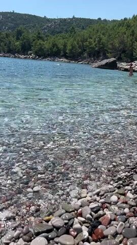 The video shows a clear, shallow body of water with a pebble-strewn seabed. The water is calm, with small waves gently lapping at the shore. In the distance, a person can be seen swimming or wading in the water. Lush green trees and hills form the backdrop, with a clear blue sky overhead. The scene appears to be a tranquil coastal environment, possibly a secluded beach.