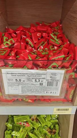 The video shows a close-up view of shelves filled with various types of wrapped candies in a store setting. The first shelf displays an abundance of red-wrapped candies with green accents, appearing to be a bulk bin. A price label is visible on a clear plastic container within this bin. The second shelf below contains green-wrapped candies, also in a bulk bin. The third shelf shows a bulk bin filled with yellow-wrapped candies, some of which have images of fruit on their wrappers. A price tag of 54.99 is visible below this shelf. The video then pans across other shelves showcasing different colored candies. One section has blue-wrapped candies, another has dark red or brown-wrapped candies, and a third has white and blue-wrapped candies. The bottom right section shows a bulk bin filled with orange-wrapped candies. Another section displays candies with purple and green wrappers. Finally, the bottom left section contains what appears to be Snickers bars, and the bottom right section shows more brown or copper-colored wrapped candies. The lighting suggests an indoor retail environment. There are no people clearly visible in the frame.
