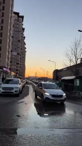The video shows a wet city street during a bright sunrise. The sky is a gradient of blue to orange. A white SUV is parked on the left side of the street, followed by other parked cars. A white and yellow taxi drives past on the right. The street is lined with modern buildings. The asphalt is reflective with puddles of water.  Bare trees and streetlights are visible along the right side of the street. Industrial structures are visible in the background.