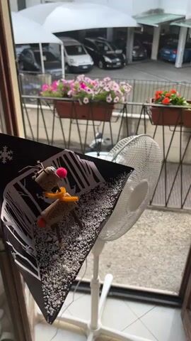 The media file is a close-up shot of a diorama featuring two small reindeer figures made from wine corks. The figures are decorated with pipe cleaners and pom-poms, and stand on a base of white gravel, which is meant to represent snow. The diorama is set against a backdrop of black and white cut-out trees, suggesting a winter forest scene. A white oscillating fan is positioned behind the diorama, partially obscuring the view.

Beyond the fan and diorama, a balcony railing is visible, adorned with planters filled with vibrant pink and orange flowers. The background shows a row of cars parked under white canopies, indicating an outdoor setting, likely a parking area or street. The lighting suggests it is daytime, possibly late morning or early afternoon, given the brightness and lack of strong shadows. The overall scene evokes a charming, handmade craft project, perhaps a holiday decoration, presented outdoors. The location context identifies Varese, Italy. There is no visible text in the media file.