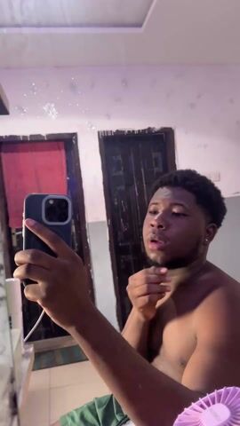 A young, shirtless Black man is taking a selfie in front of a bathroom mirror. He is holding an iPhone in his left hand and looking at the phone screen. The background includes a white and beige wall with a ceiling light, a red door, and a brown wooden door. A small purple electric fan is partially visible in the bottom right corner. The mirror has water spots on it. The man has a short beard and curly hair. He is wearing an earring in his left ear. He appears to be indoors.