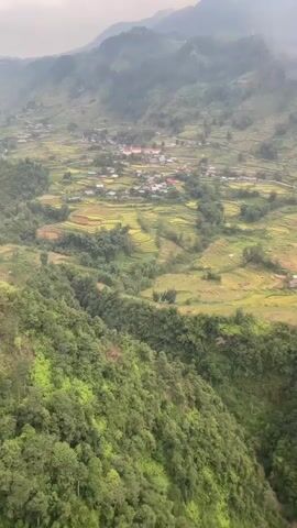 The video pans over a lush, mountainous landscape covered in dense green trees. Below, a village is nestled in a valley amidst terraced rice fields, some appearing golden and ready for harvest. The sky is overcast with clouds, and the overall scene suggests a rural, agricultural environment in a hilly or mountainous region. There are no people or specific objects clearly visible within the frame.