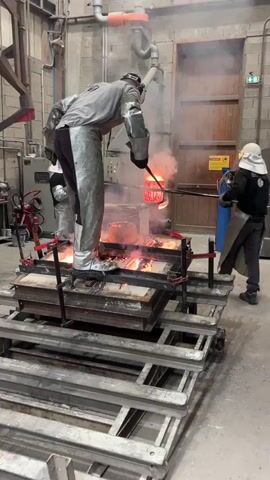 The video depicts a metal casting operation in an industrial foundry located in Saint-Rémy-lès-Chevreuse, France. Three individuals, all clad in extensive personal protective equipment (PPE) including heat-resistant silver suits, chaps, gloves, and face shields, are actively involved in the process.

In the foreground, a worker stands on an elevated metal platform, carefully pouring intensely glowing orange molten metal from a crucible, held by a long-handled tool, into a mold situated on the platform. Several molds already filled with molten metal on the platform are brightly incandescent, emitting a vivid orange and red glow.

To the right of the main worker, a second individual, wearing a dark jacket, silver apron, and a white hood/face shield, assists with another long tool. A third worker is partially visible on the left, also in protective gear. Smoke or steam rises visibly from the molten metal and the surrounding area, indicating high temperatures.

The background shows an industrial workshop environment with brick walls, metal structures, white pipes, and machinery, likely part of a furnace system where crucibles are heated. A red fire extinguisher is mounted on the left wall. The scene illustrates a precise and hazardous industrial process demanding stringent safety measures due to the extreme heat involved. No discernible text is legible on any signs or equipment within the frame.