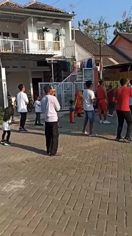 The video shows a group of people gathered on a paved street in front of houses. Some are standing still, while others appear to be engaged in some form of coordinated activity, possibly exercise or a dance, led by individuals in the foreground. The people are dressed in casual clothing, with some wearing white shirts and others red. The environment is outdoors, with clear blue sky visible. There are houses in the background, some with multiple stories. A bicycle is leaning against one of the houses, and clothes are hanging on a line. The lighting suggests it is daytime, possibly morning or afternoon. The overall scene depicts a community gathering or event taking place in a residential area.